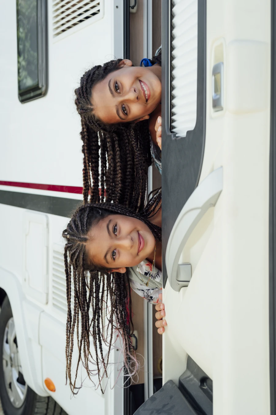 Deux jeunes filles souriantes aux tresses longues regardant par la porte d’une roulotte blanche, symbolisant le plaisir du voyage en famille en VR.