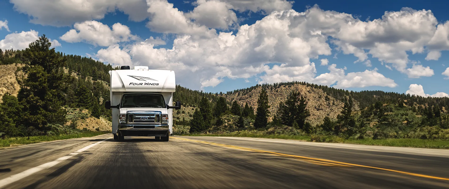 Motorisé Thor Four Winds roulant sur une route de montagne bordée de pins sous un ciel bleu, mettant en valeur la liberté du voyage en VR.